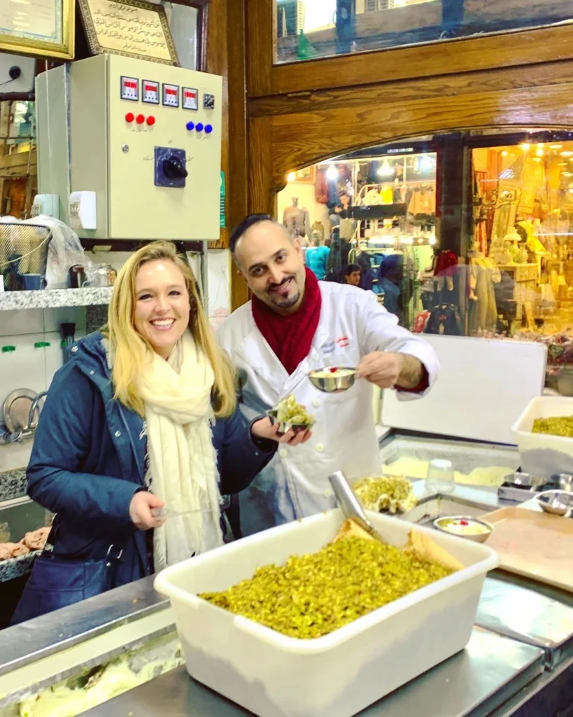 A smiling woman stands behind the counter at the famous Bakdash ice cream parlor in Damascus, holding a bowl of traditional ice cream, while a staff member in a white uniform prepares another serving in front of a large container of crushed pistachios.