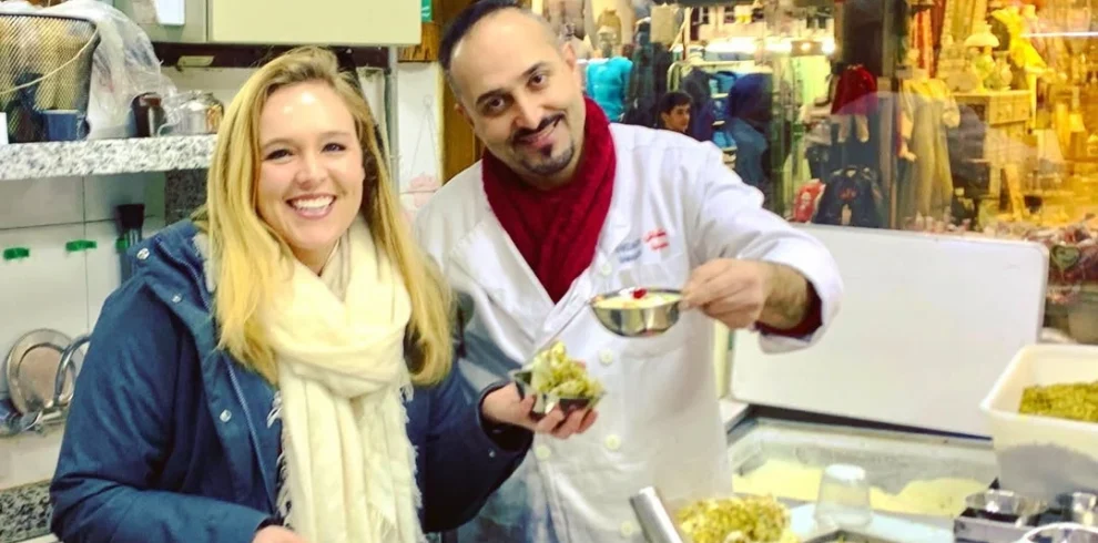 A smiling woman stands behind the counter at the famous Bakdash ice cream parlor in Damascus, holding a bowl of traditional ice cream, while a staff member in a white uniform prepares another serving in front of a large container of crushed pistachios.