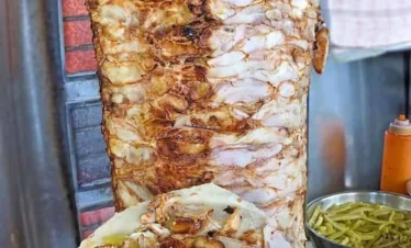 A close-up of a large, vertically roasting spit of seasoned chicken shawarma with a hand holding an open flatbread filled with shaved meat, garlic sauce, and pickles in a local Syrian eatery.