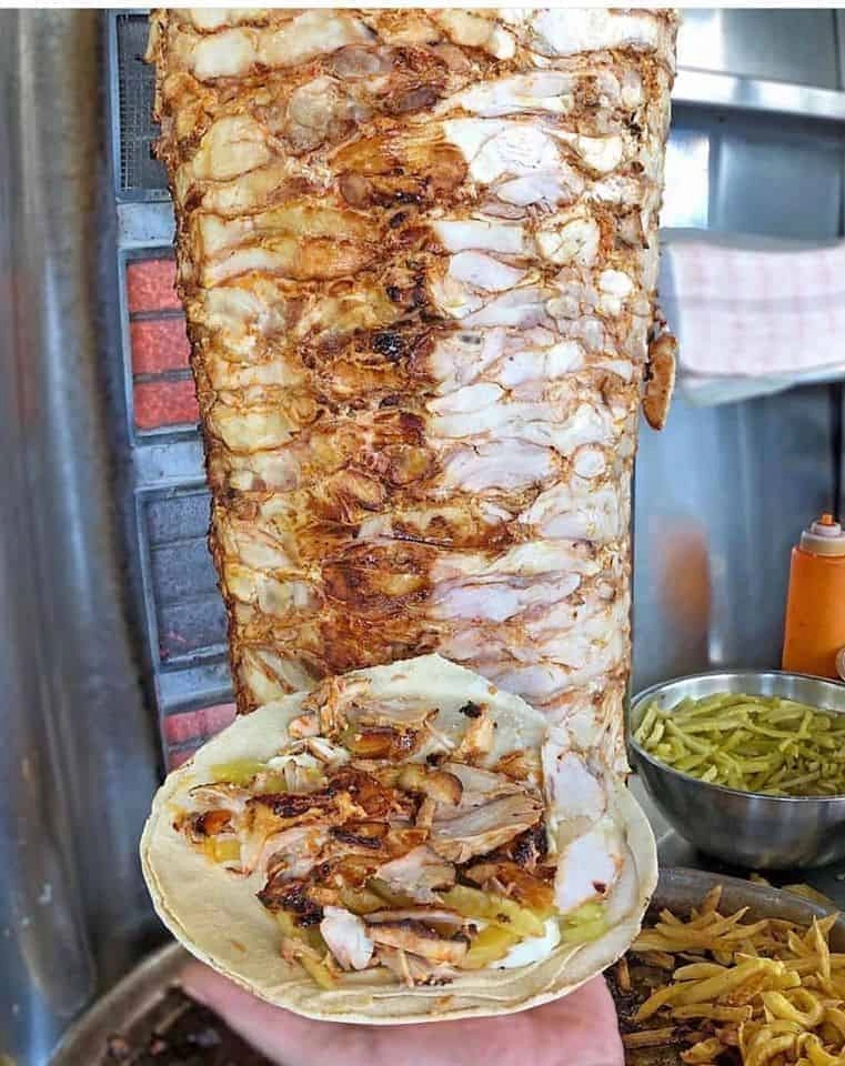A close-up of a large, vertically roasting spit of seasoned chicken shawarma with a hand holding an open flatbread filled with shaved meat, garlic sauce, and pickles in a local Syrian eatery.