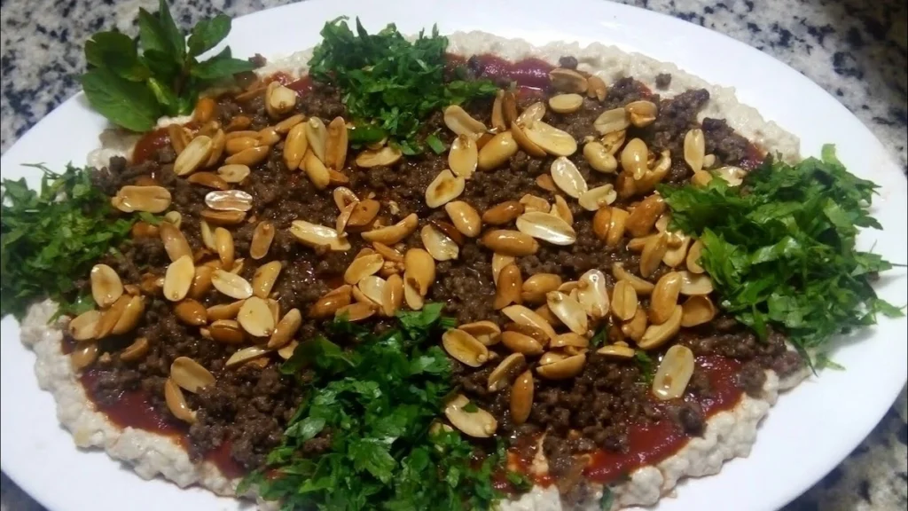 A large oval serving platter featuring Batrash, a base of creamy eggplant spread topped with a layer of red tomato sauce, seasoned ground meat, a generous handful of roasted peanuts, and fresh green parsley garnish.