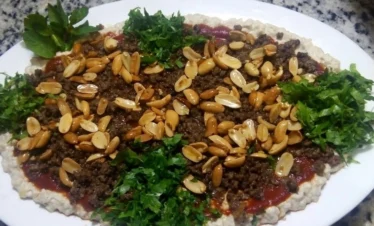 A large oval serving platter featuring Batrash, a base of creamy eggplant spread topped with a layer of red tomato sauce, seasoned ground meat, a generous handful of roasted peanuts, and fresh green parsley garnish.