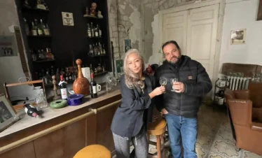 A man and a woman stand behind a vintage wooden bar holding wine glasses for a toast, set in a rustic room with exposed stone walls, traditional patterned tile floors, and a back bar display of assorted bottles.