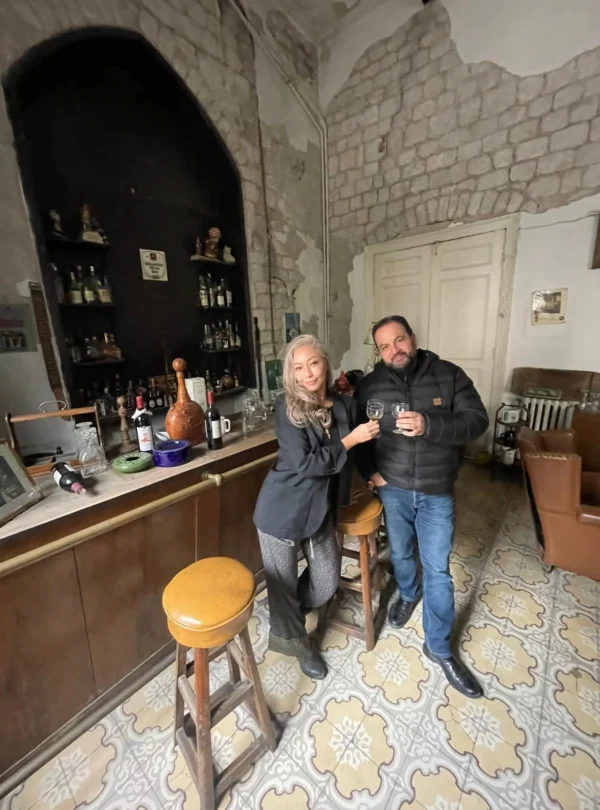 A man and a woman stand behind a vintage wooden bar holding wine glasses for a toast, set in a rustic room with exposed stone walls, traditional patterned tile floors, and a back bar display of assorted bottles.