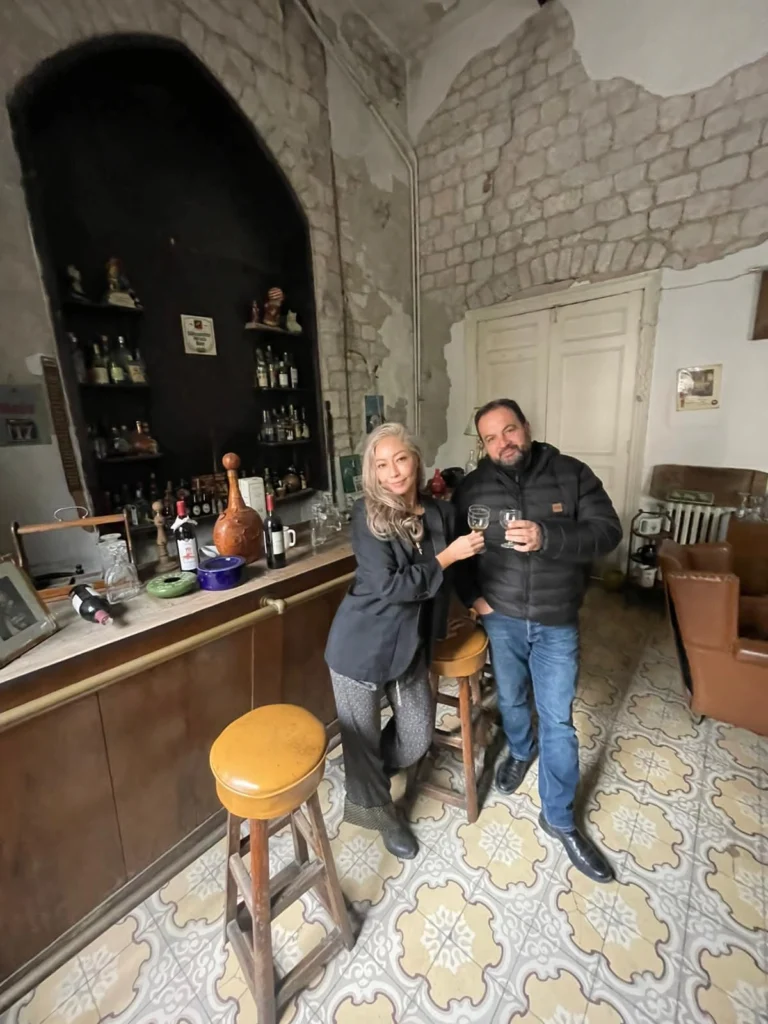 A man and a woman stand behind a vintage wooden bar holding wine glasses for a toast, set in a rustic room with exposed stone walls, traditional patterned tile floors, and a back bar display of assorted bottles.