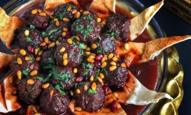 A glass bowl filled with dark, glistening meatball kebabs in a deep red cherry sauce, garnished with toasted pine nuts and pomegranate seeds, arranged with decorative triangles of crispy fried pita bread.
