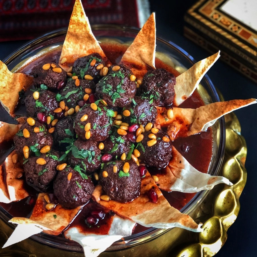A glass bowl filled with dark, glistening meatball kebabs in a deep red cherry sauce, garnished with toasted pine nuts and pomegranate seeds, arranged with decorative triangles of crispy fried pita bread.