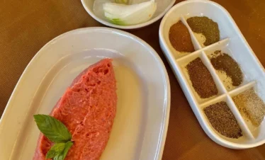 A top-down view of a plate of Kibbeh Nayyeh—finely ground raw lamb mixed with bulgur—garnished with fresh mint and served with sliced onions on ice and a side tray of six different aromatic spices.