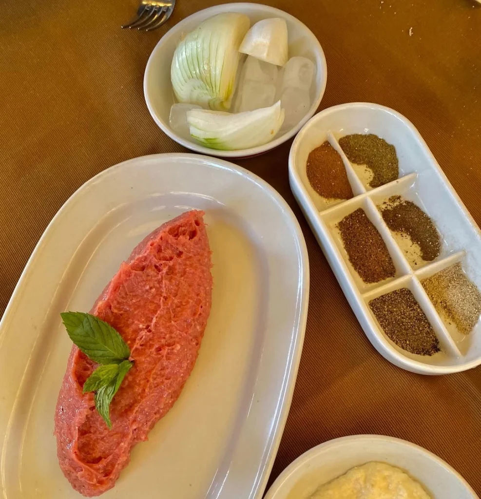 A top-down view of a plate of Kibbeh Nayyeh—finely ground raw lamb mixed with bulgur—garnished with fresh mint and served with sliced onions on ice and a side tray of six different aromatic spices.