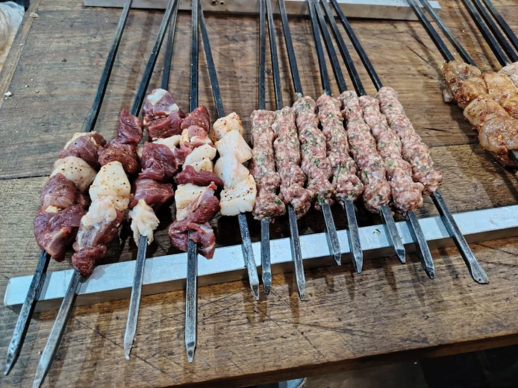 A top-down view of several long metal skewers on a wooden table, some loaded with chunks of raw lamb and white fat, and others with seasoned ground meat kebabs, ready for grilling.