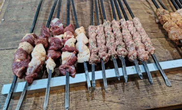 A top-down view of several long metal skewers on a wooden table, some loaded with chunks of raw lamb and white fat, and others with seasoned ground meat kebabs, ready for grilling.