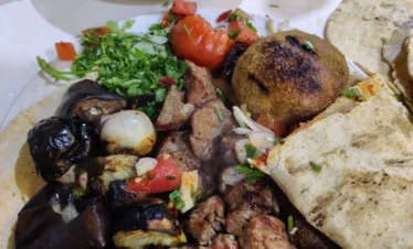 A close-up, top-down view of a loaded dinner plate featuring grilled lamb chunks, minced meat kebabs, a large charred grilled kibbeh, and roasted vegetables, served with a side of creamy hummus and fresh flatbread.