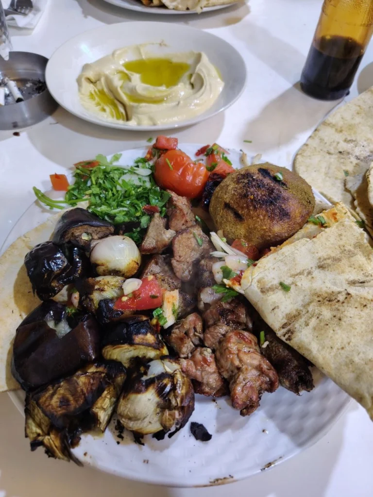 A close-up, top-down view of a loaded dinner plate featuring grilled lamb chunks, minced meat kebabs, a large charred grilled kibbeh, and roasted vegetables, served with a side of creamy hummus and fresh flatbread.