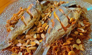 A close-up view of a large platter featuring two whole fried fish served over a generous mound of dark, caramelized onion-spiced rice, topped with a scattering of toasted almonds and cashews.