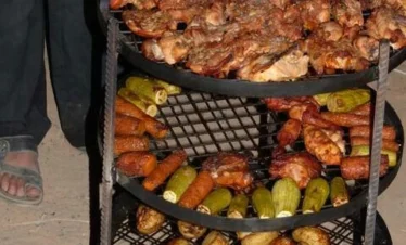 A three-tiered metal grilling rack being lifted from an underground oven, with the top tier filled with roasted chicken and the middle and bottom tiers loaded with grilled zucchini, carrots, and potatoes.