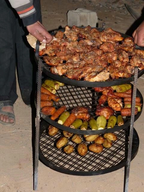 A three-tiered metal grilling rack being lifted from an underground oven, with the top tier filled with roasted chicken and the middle and bottom tiers loaded with grilled zucchini, carrots, and potatoes.