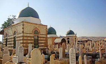 A view of the historic Bab al-Saghir cemetery in Damascus, featuring several stone mausoleums with prominent green domes and striped masonry, surrounded by numerous white carved headstones under a clear blue sky.