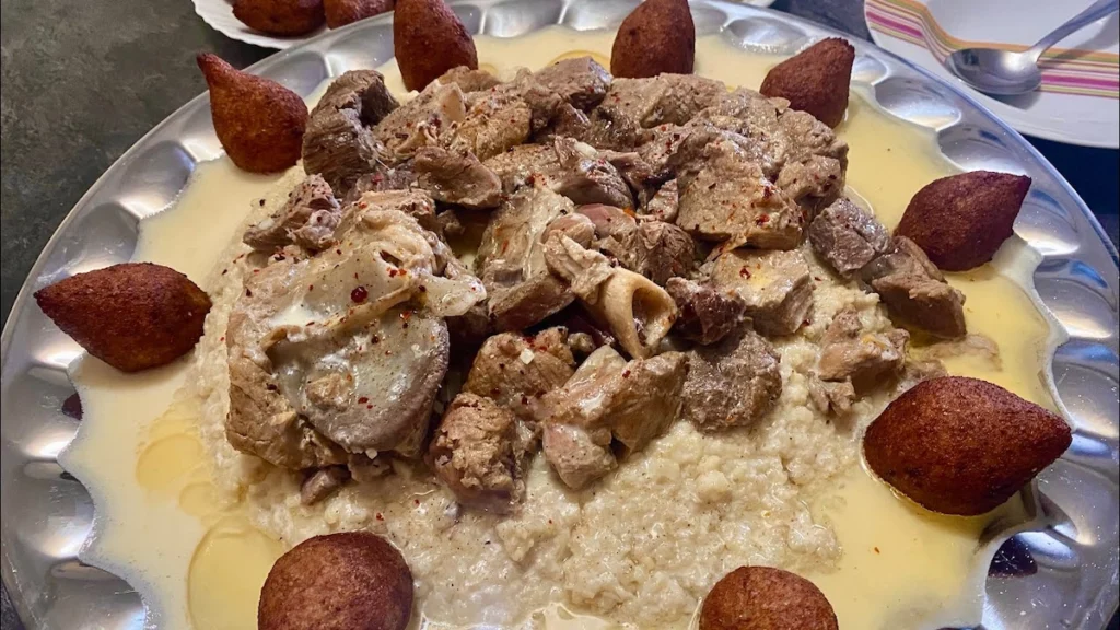 A large communal silver platter of Mlehi, featuring a bed of bulgur wheat topped with tender chunks of meat and surrounded by golden-fried kibbeh balls, all drizzled with a rich, creamy yogurt-based sauce.