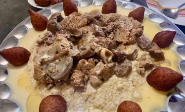 A large communal silver platter of Mlehi, featuring a bed of bulgur wheat topped with tender chunks of meat and surrounded by golden-fried kibbeh balls, all drizzled with a rich, creamy yogurt-based sauce.