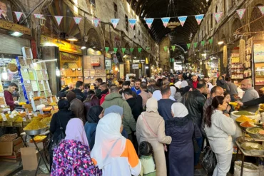 A high-angle view of a crowded, vaulted alleyway in the historic Al-Hamidiyah Souq in Damascus, with vendors selling colorful spices and sweets under hanging festive banners.
