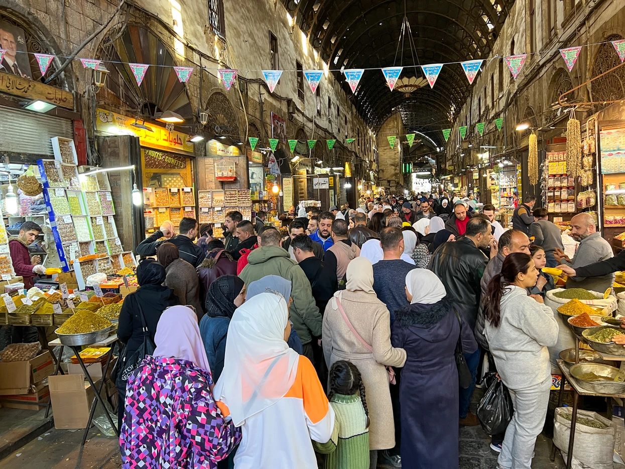 A high-angle view of a crowded, vaulted alleyway in the historic Al-Hamidiyah Souq in Damascus, with vendors selling colorful spices and sweets under hanging festive banners.