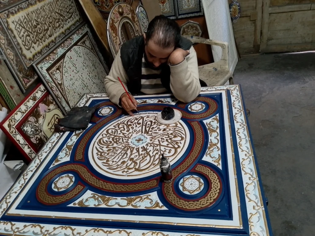 A skilled Syrian craftsman in his workshop meticulously hand-painting intricate Arabic calligraphy and geometric patterns onto a large, decorative wooden panel in Damascus.