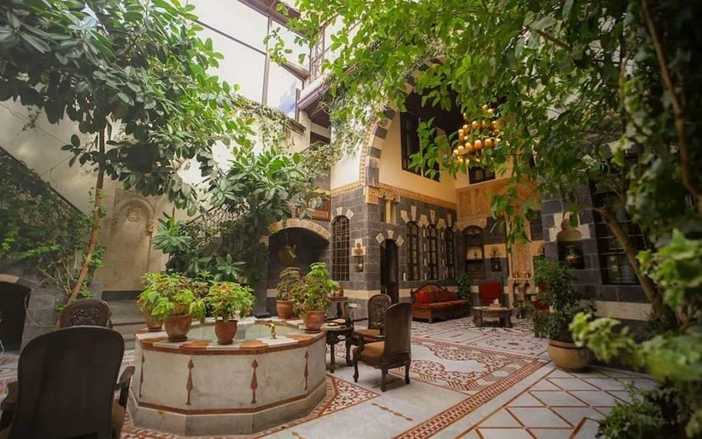 The traditional interior courtyard of Beit Al-Wali boutique hotel in Old Damascus, featuring a central marble fountain, lush greenery, and intricate stone mosaic flooring.