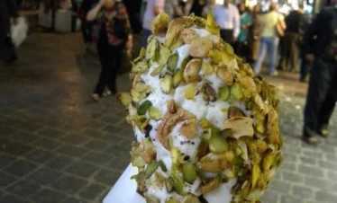 A close-up of a hand holding a waffle cone piled high with stretchy Damascene ice cream (Booza), heavily coated in crushed bright green pistachios and cashews, with the blurred lights of Souq Al-Hamidiyah in the background.