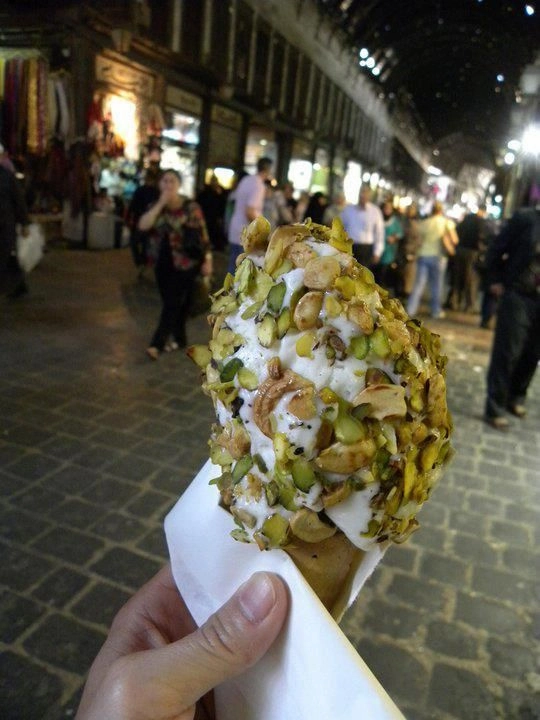 A close-up of a hand holding a waffle cone piled high with stretchy Damascene ice cream (Booza), heavily coated in crushed bright green pistachios and cashews, with the blurred lights of Souq Al-Hamidiyah in the background.
