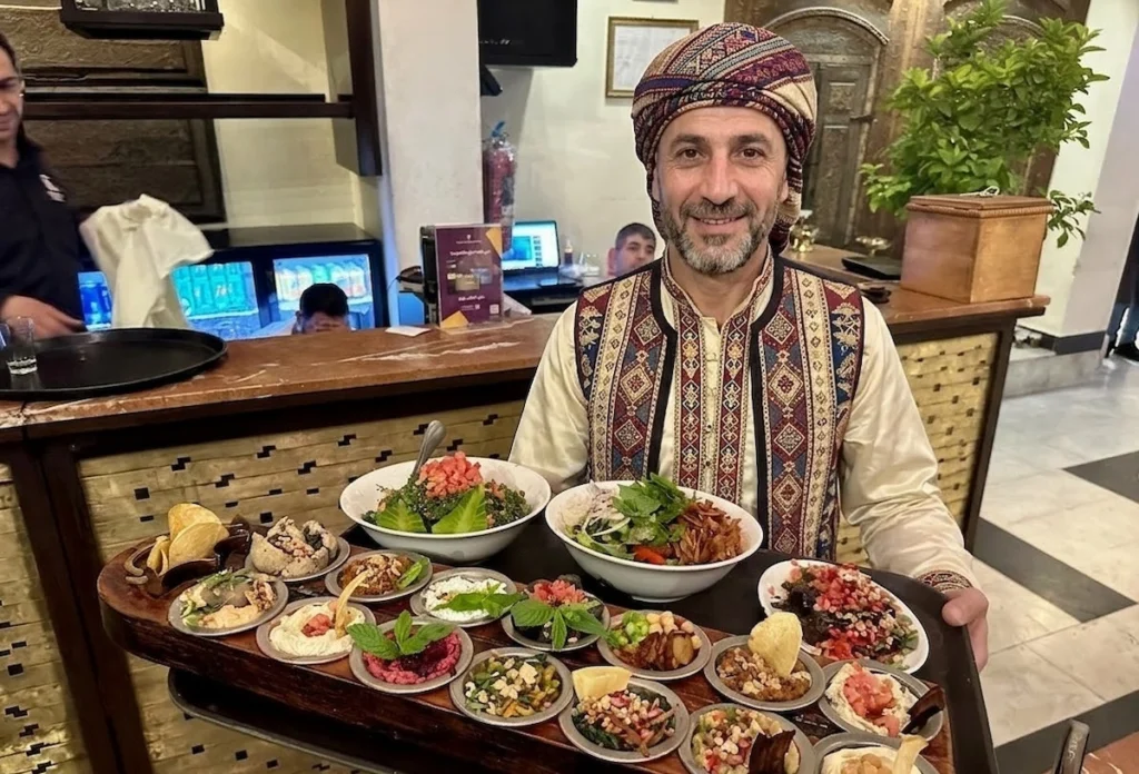 A smiling man in traditional Damascene attire holding a large wooden tray filled with a variety of Syrian mezze dishes, including hummus, tabbouleh, and fattoush, in a Damascus restaurant.
