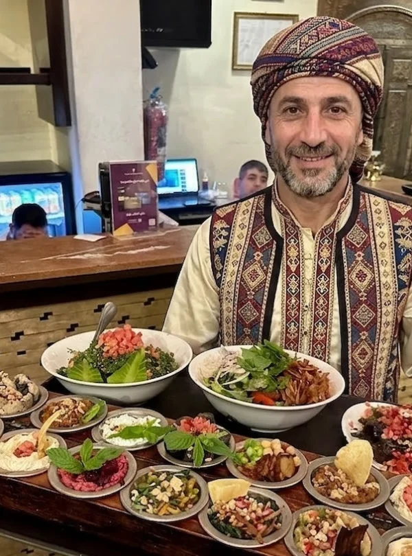 A smiling man in traditional Damascene attire holding a large wooden tray filled with a variety of Syrian mezze dishes, including hummus, tabbouleh, and fattoush, in a Damascus restaurant.