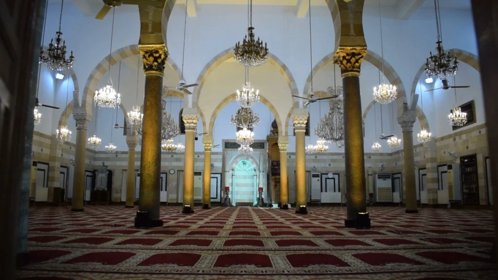 The expansive prayer hall of the Ibn Arabi Mosque in Damascus, featuring high arched ceilings, numerous ornate crystal chandeliers hanging from long cords, and prominent stone columns with golden capitals atop red patterned carpets.