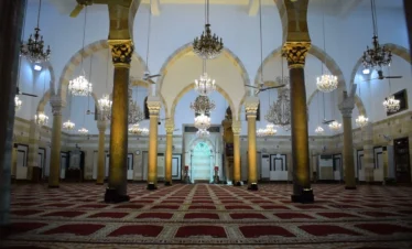 The expansive prayer hall of the Ibn Arabi Mosque in Damascus, featuring high arched ceilings, numerous ornate crystal chandeliers hanging from long cords, and prominent stone columns with golden capitals atop red patterned carpets.