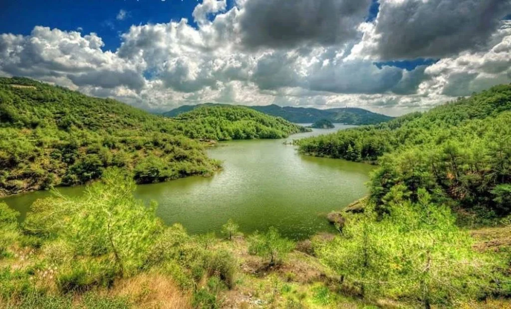 An elevated view of the lush, forested valley surrounding the tranquil, deep green waters of the Wadi Qandil lake near Lattakia, Syria, with rolling hills under dramatic cloudy skies.