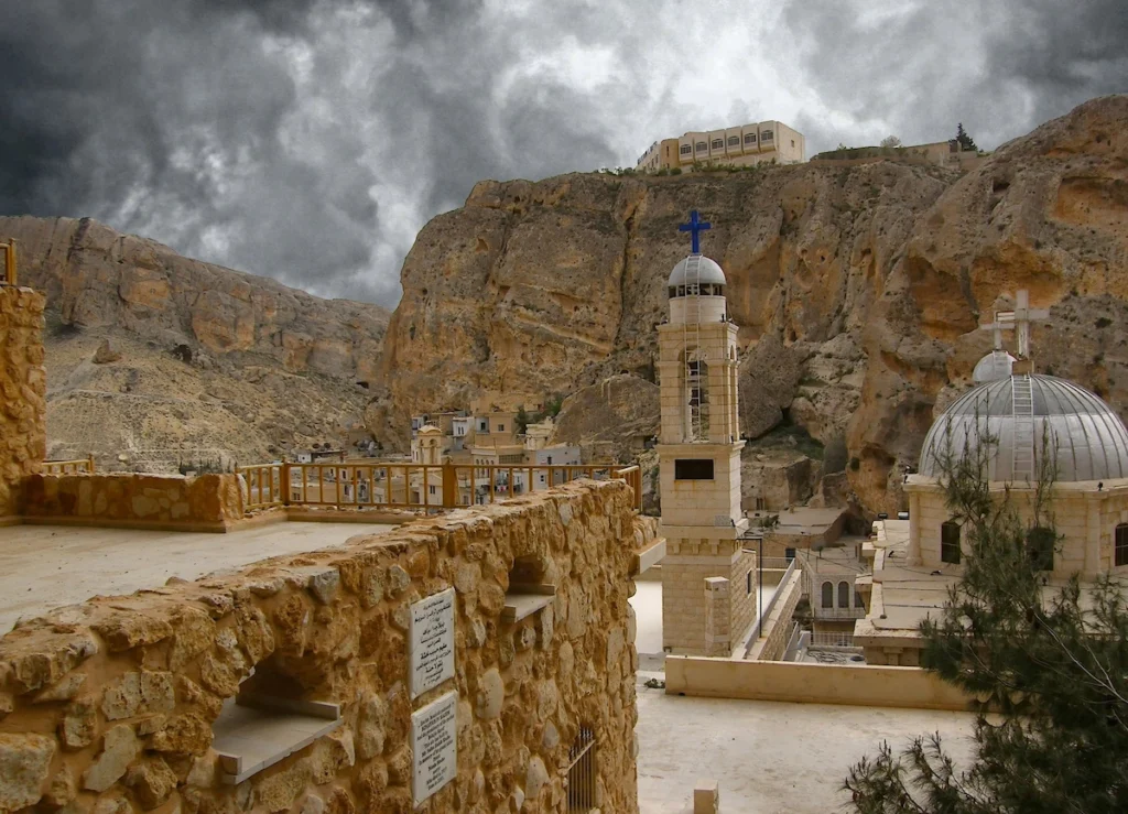 An elevated view of the historic Aramaic village of Maaloula, Syria, featuring ancient cliff-carved architecture, a church belfry with a blue cross, a domed building with a white cross, and a building on a high peak under dramatic cloudy skies.