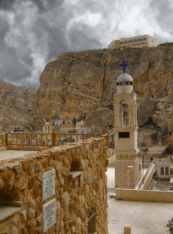An elevated view of the historic Aramaic village of Maaloula, Syria, featuring ancient cliff-carved architecture, a church belfry with a blue cross, a domed building with a white cross, and a building on a high peak under dramatic cloudy skies.