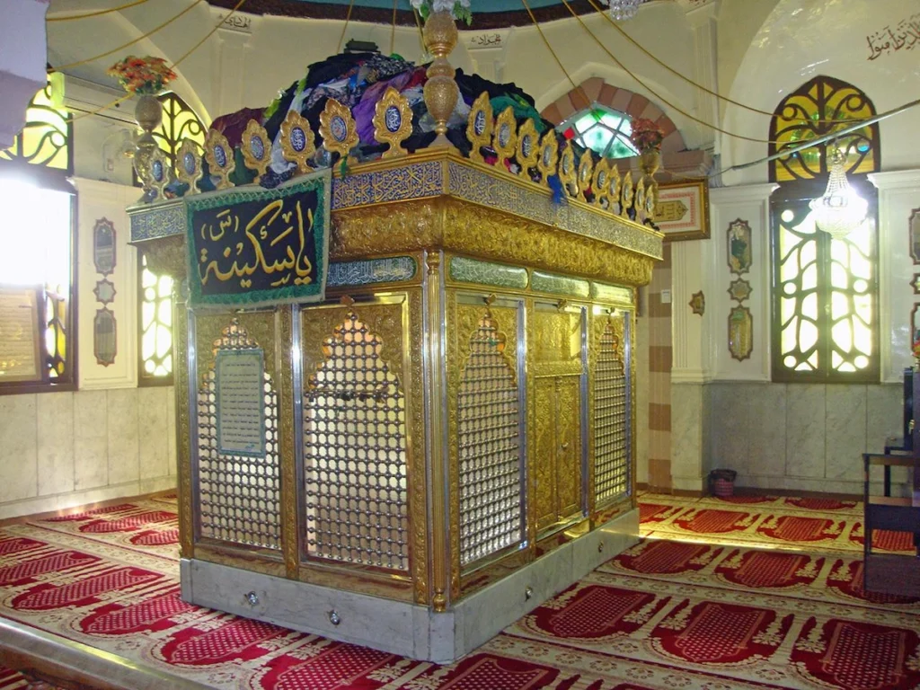 A golden, intricately carved metal shrine (zarih) with a green and gold banner, situated inside a prayer hall with red patterned carpets and arched windows at the Bab al-Saghir cemetery in Damascus.