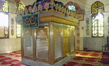 A golden, intricately carved metal shrine (zarih) with a green and gold banner, situated inside a prayer hall with red patterned carpets and arched windows at the Bab al-Saghir cemetery in Damascus.