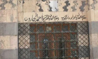 A close-up of the exterior stone wall of the Maqam of Sheikh Arslan in Damascus, featuring traditional black and white ablaq masonry, intricate stone carvings, and elegant Arabic calligraphy inscribed above a barred window.