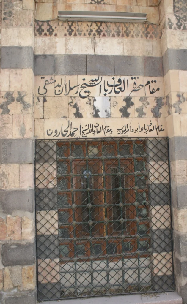 A close-up of the exterior stone wall of the Maqam of Sheikh Arslan in Damascus, featuring traditional black and white ablaq masonry, intricate stone carvings, and elegant Arabic calligraphy inscribed above a barred window.