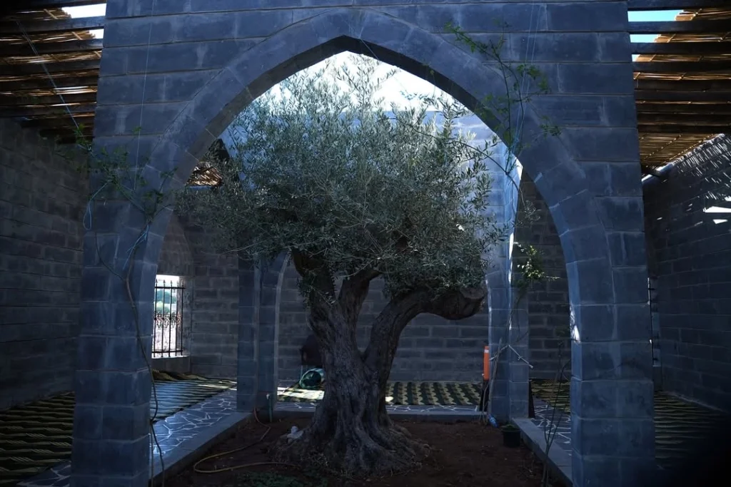 A view of the historic Mausoleum of Imam al-Nawawi in the city of Nawa, featuring a traditional stone structure with a small dome, set against a backdrop of modern residential buildings and green trees.