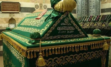 The ornate tomb of Saladin in Damascus, featuring a green velvet cover with intricate gold Arabic calligraphy and tassels, set against a background of historic patterned stone walls.