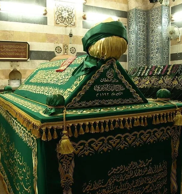 The ornate tomb of Saladin in Damascus, featuring a green velvet cover with intricate gold Arabic calligraphy and tassels, set against a background of historic patterned stone walls.