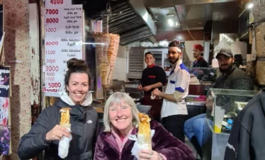 Two smiling female tourists standing in front of a traditional Syrian shawarma shop in Old Damascus, each holding a wrapped chicken shawarma sandwich while staff prepare food in the background.