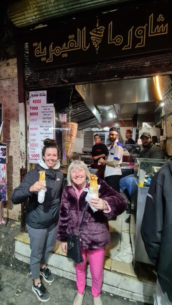 Two smiling female tourists standing in front of a traditional Syrian shawarma shop in Old Damascus, each holding a wrapped chicken shawarma sandwich while staff prepare food in the background.