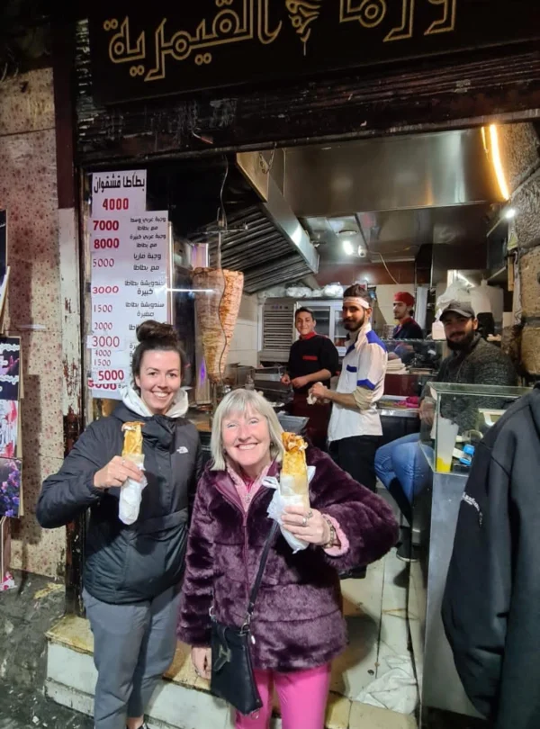 Two smiling female tourists standing in front of a traditional Syrian shawarma shop in Old Damascus, each holding a wrapped chicken shawarma sandwich while staff prepare food in the background.