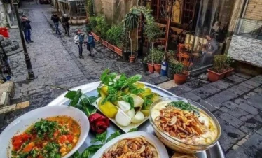 A silver tray of traditional Syrian food, including Fatteh with nuts, hummus with minced meat, and fresh greens, set against a background of a narrow cobblestone street in Old Damascus with people walking past historic stone buildings.