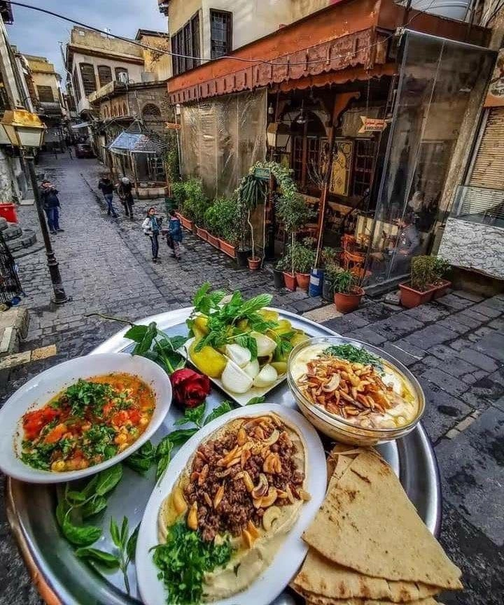 A silver tray of traditional Syrian food, including Fatteh with nuts, hummus with minced meat, and fresh greens, set against a background of a narrow cobblestone street in Old Damascus with people walking past historic stone buildings.