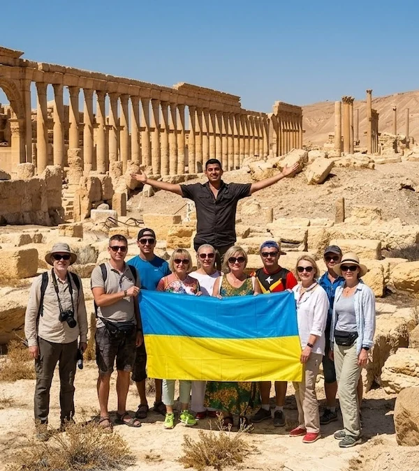 A diverse group of tourists holding a large Ukrainian flag and a man with arms outstretched pose in the center of the ancient Roman ruins of Palmyra, Syria.