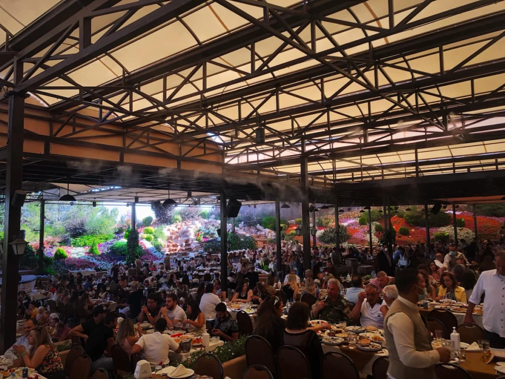 A high-angle shot of a large, crowded outdoor garden restaurant with a metal-framed canopy, misting fans, and hundreds of people dining at tables, set against a backdrop of vibrant, multicolored flower hills and rock waterfalls.